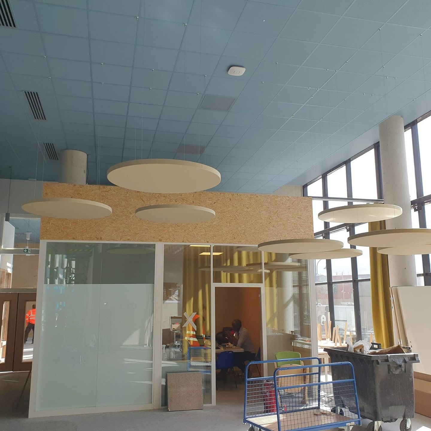 Hall vitré dans un bâtiment moderne, avec plafonds bleus et panneaux acoustiques suspendus circulaires.