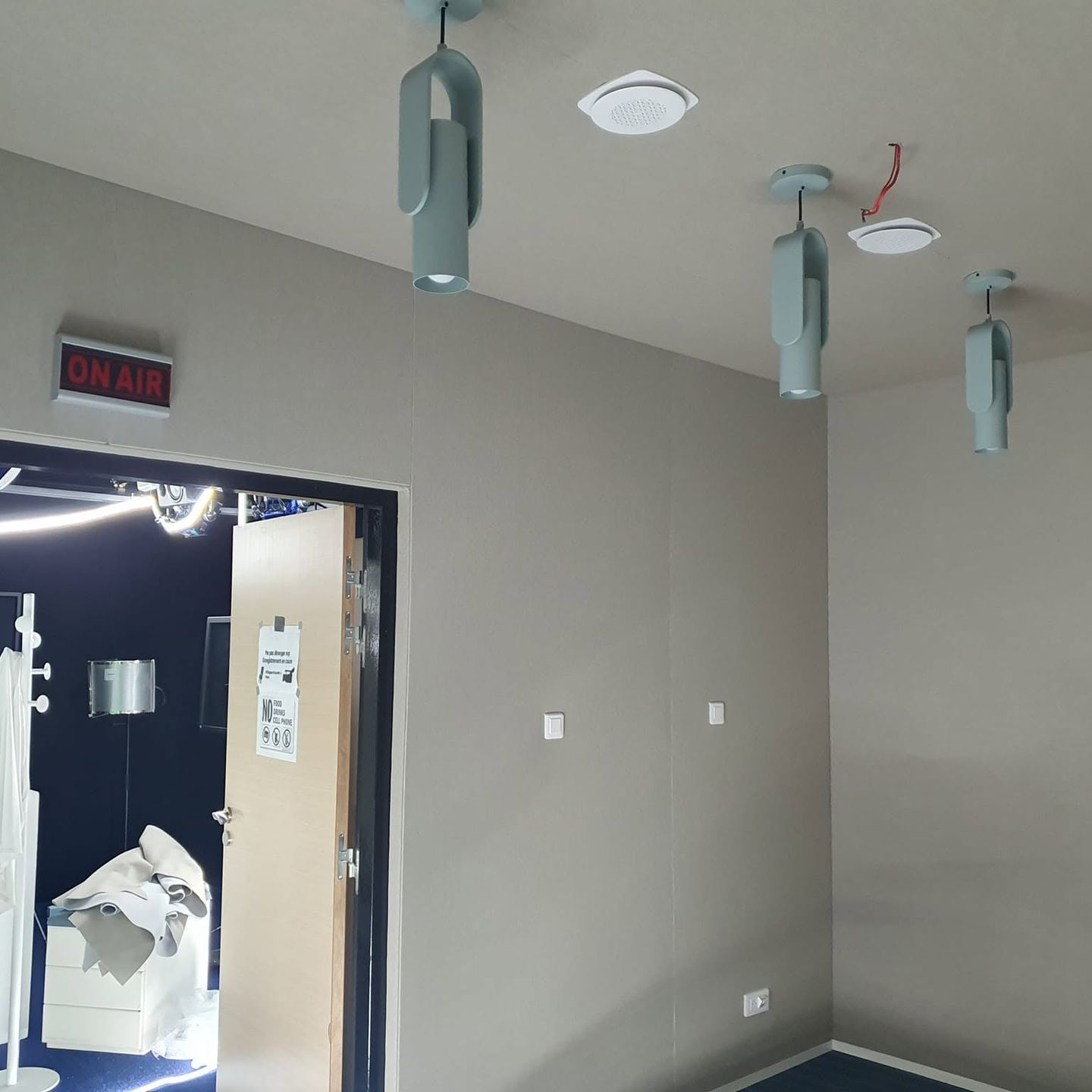 Espace intérieur en cours d’aménagement avec suspensions cylindriques au plafond et porte ouverte sur un studio d’enregistrement signalé par un panneau On Air.