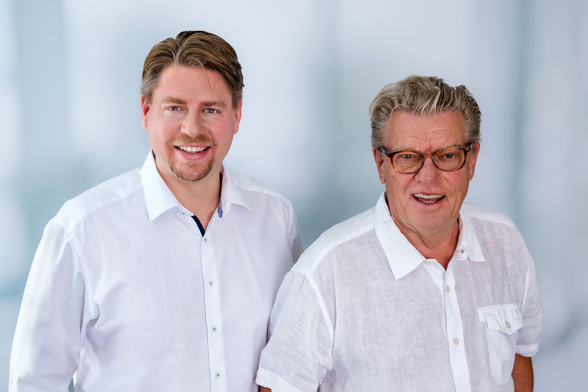 Zwei Männer stehen lächelnd nebeneinander. Einer ist jünger und trägt ein weißes Hemd, der andere ist älter und trägt eine Brille und ein weißes Leinenhemd.