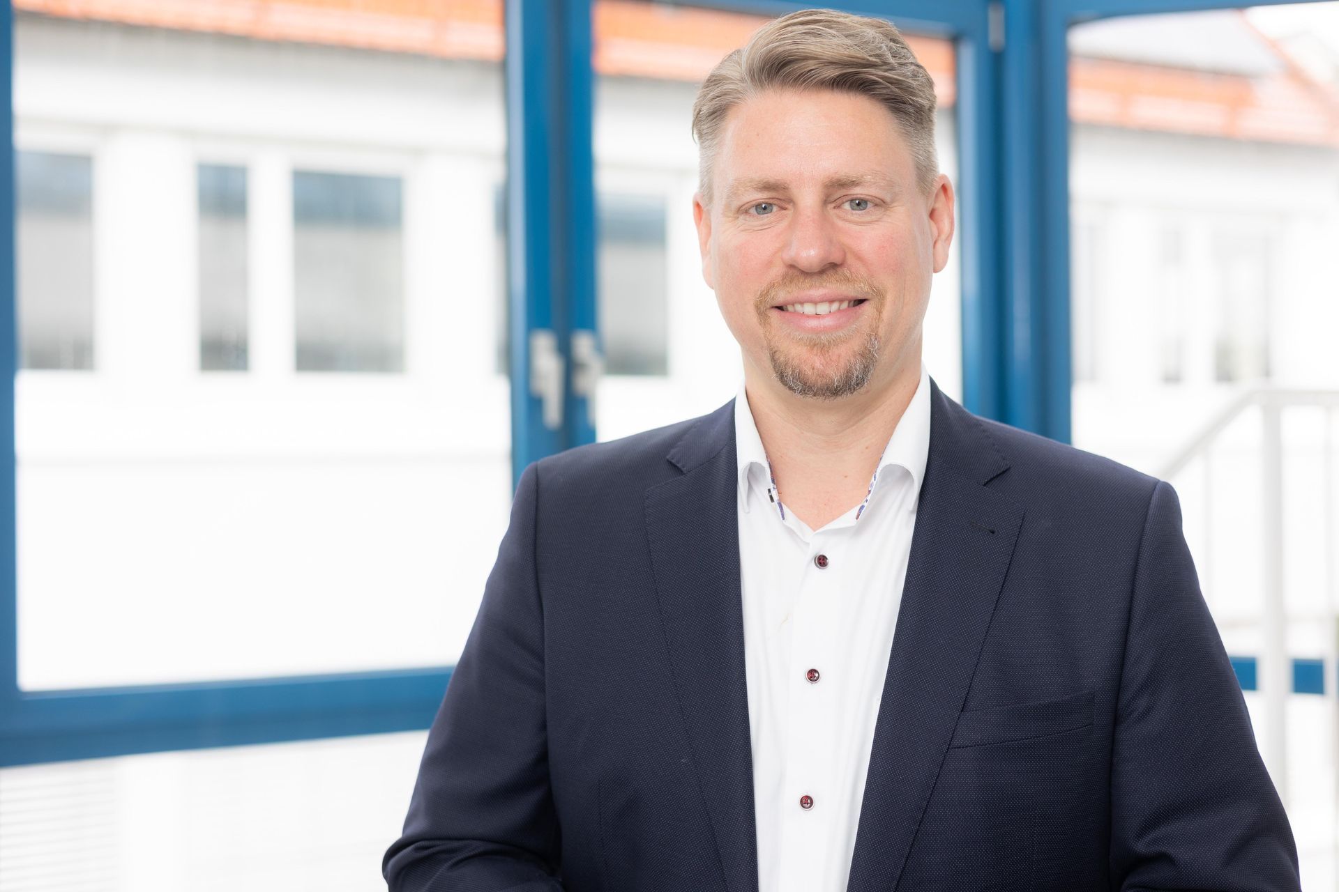 Mann im Blazer lächelt, steht neben einem Fenster mit blauem Rahmen, hellem Hintergrund.