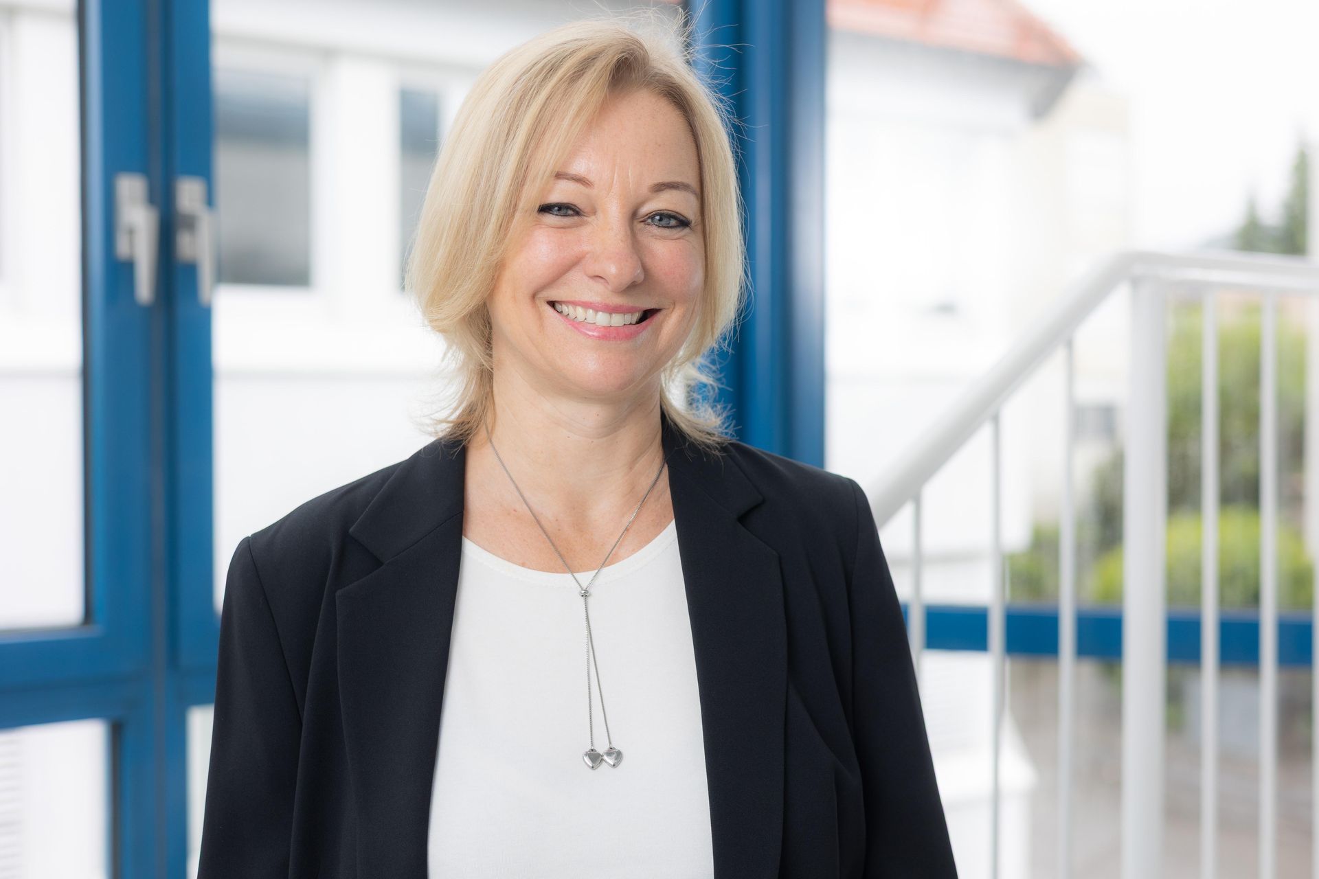 Lächelnde blonde Frau in schwarzem Blazer, weißem Hemd und Halskette in einem hellen Büroumfeld.