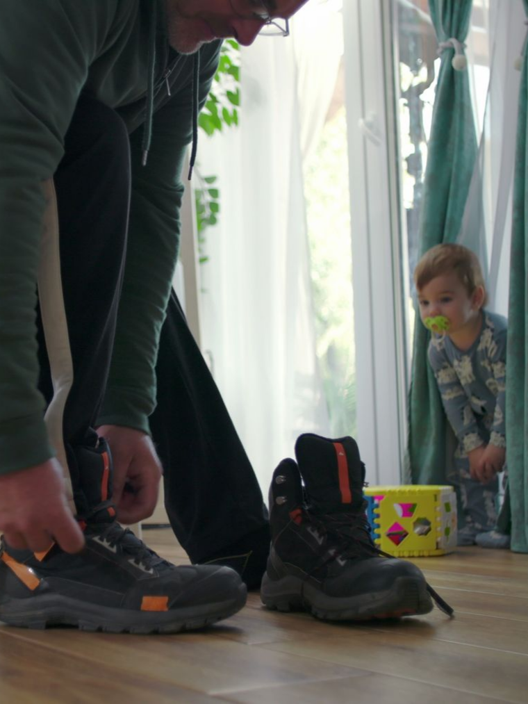 Un homme attache des bottes de travail, un enfant regarde derrière les rideaux