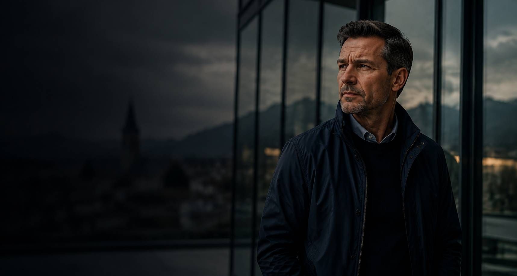 Mann in dunkler Jacke steht in der Abenddämmerung am Fenster und blickt mit ernstem Gesichtsausdruck hinaus.