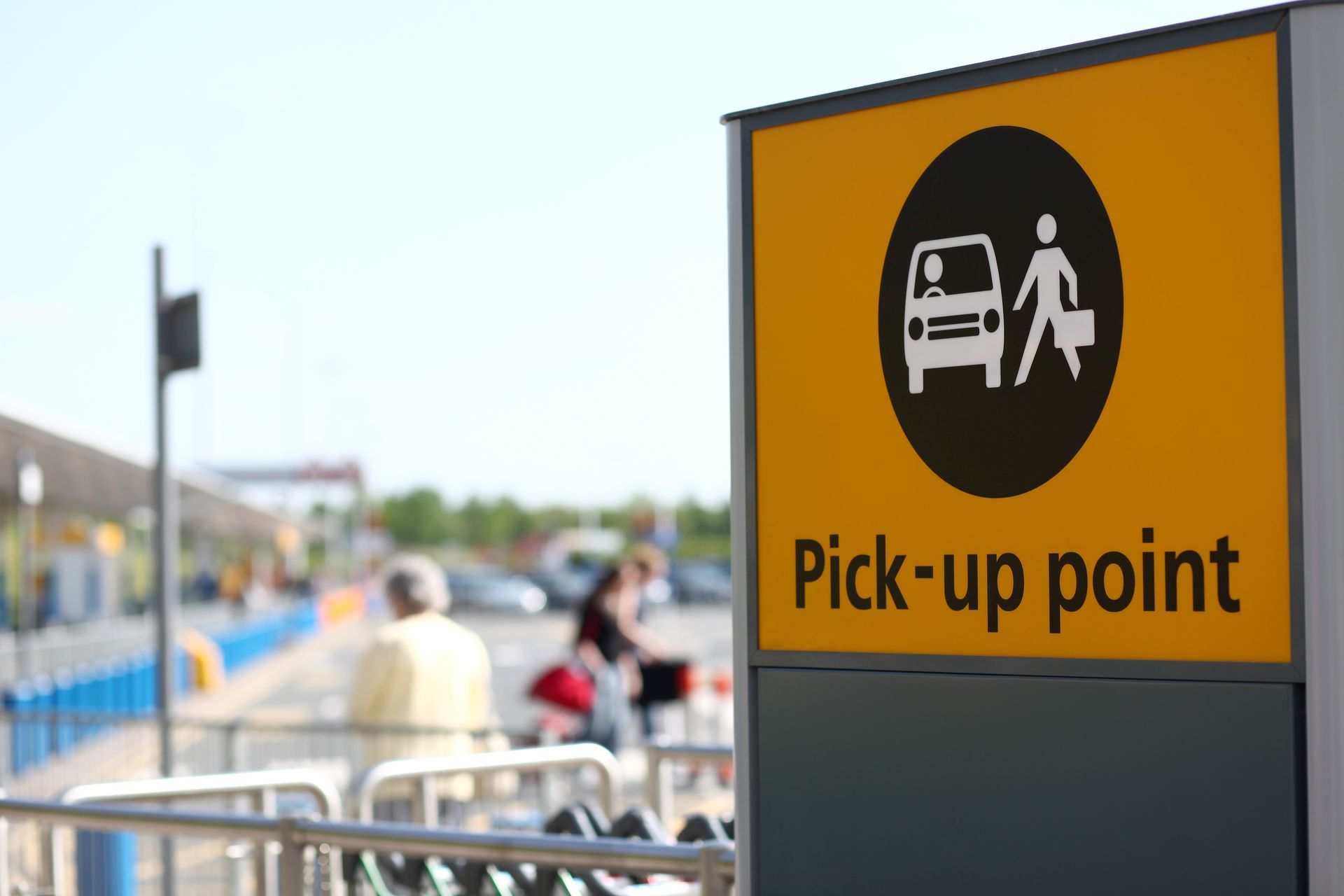Un panneau jaune point de prise en charge avec une icône représentant une voiture et un piéton dans un parking d'aéroport, avec des personnes floues en arrière-plan.