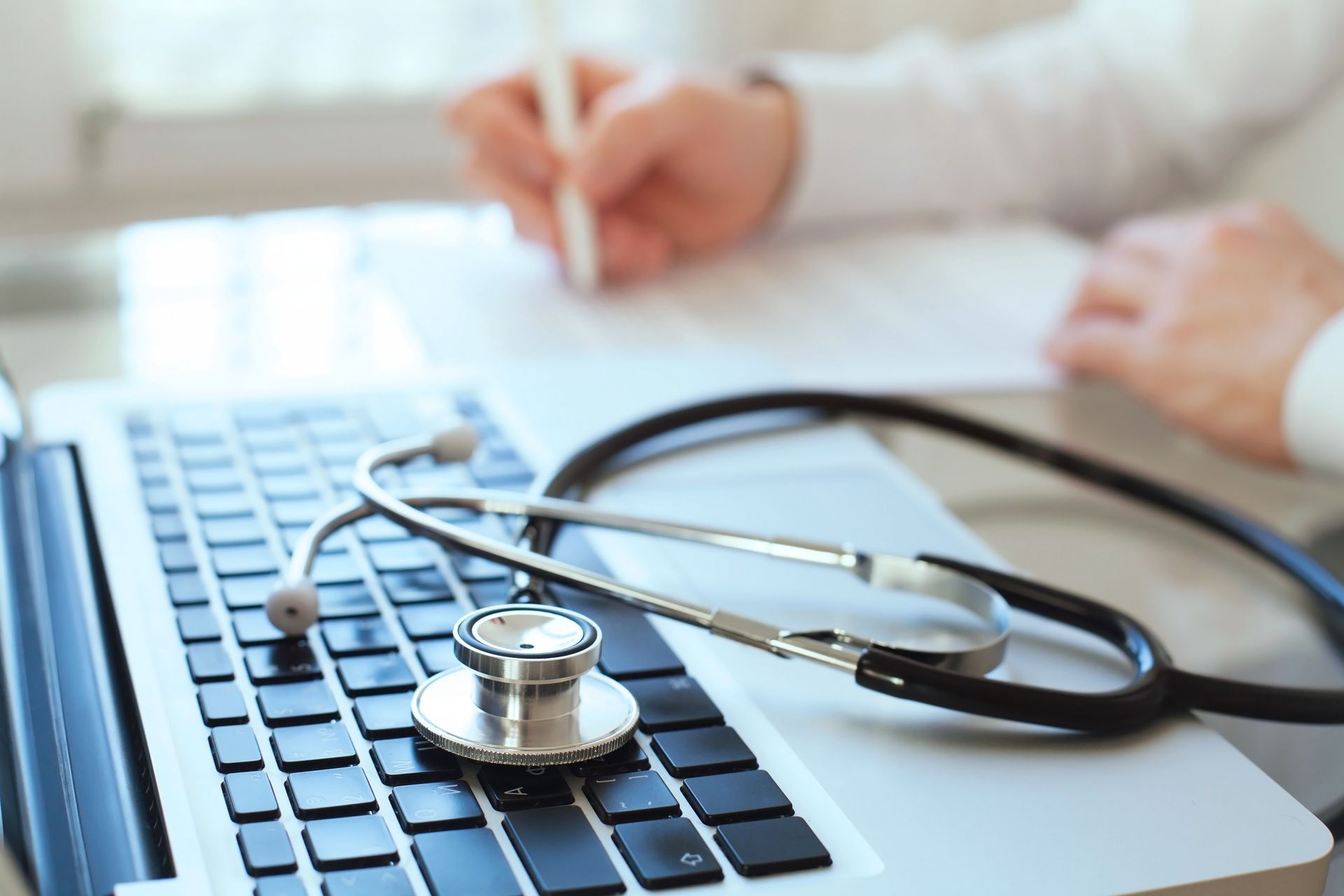 Un stéthoscope repose sur le clavier d'un ordinateur portable, tandis qu'une personne à l'arrière-plan écrit sur un document.