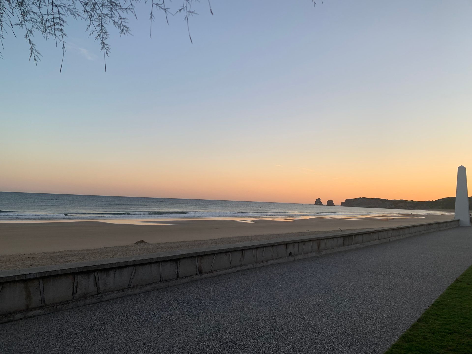 Un coucher de soleil illumine une plage de sable tranquille, avec un chemin de pierre et une colonne au premier plan.