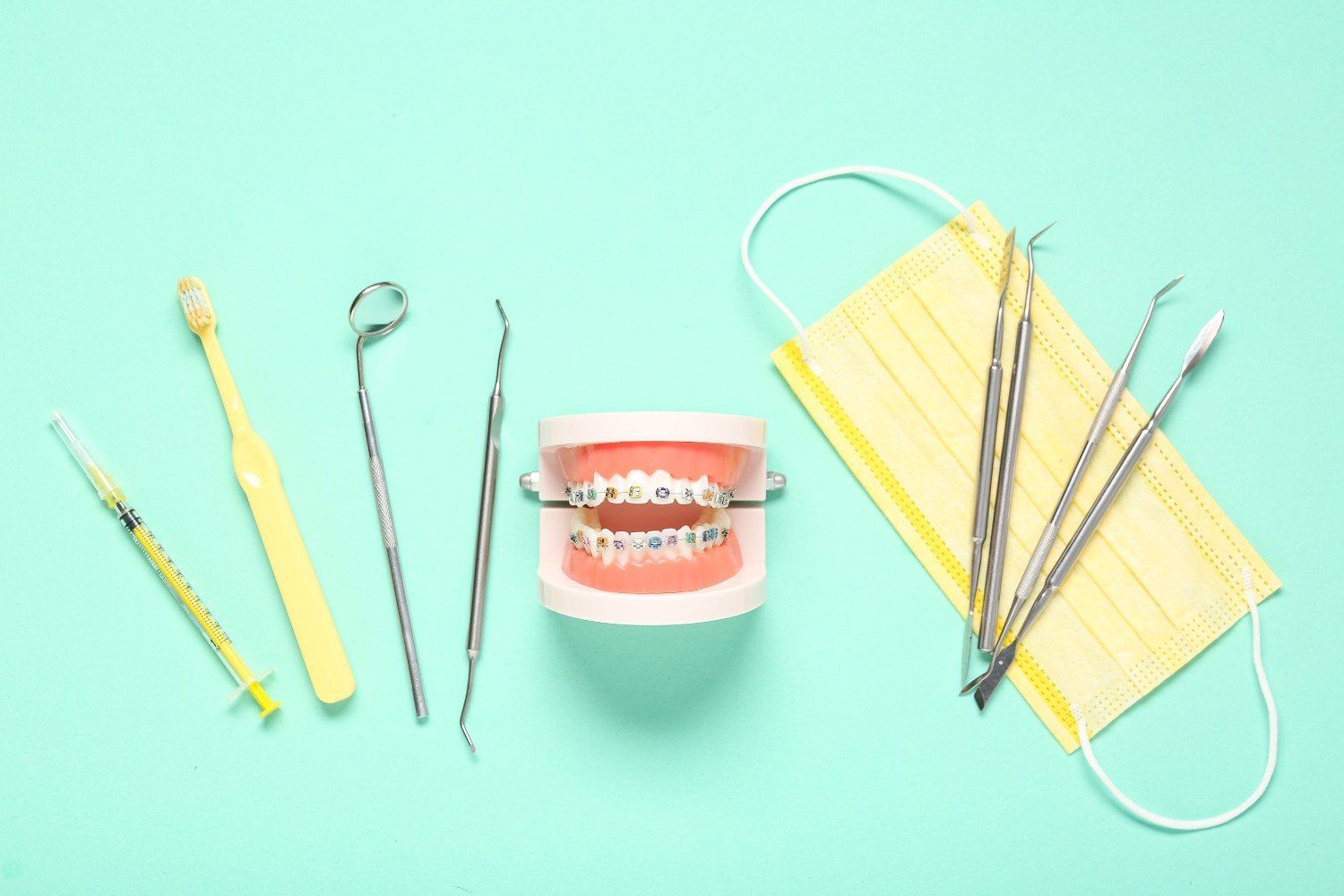 Modelo de instrumental dental y aparatos de ortodoncia sobre fondo menta con una mascarilla amarilla.