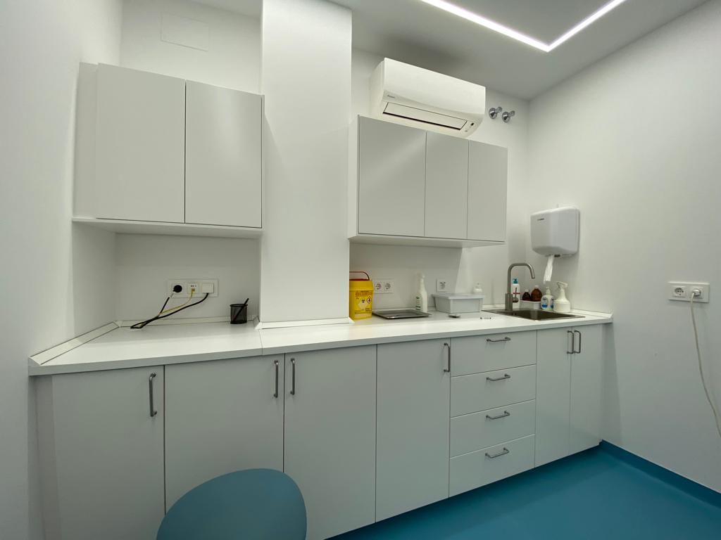 Pequeña cocina clínica blanca con armarios, fregadero, encimera, aire acondicionado de pared y suelo azul.