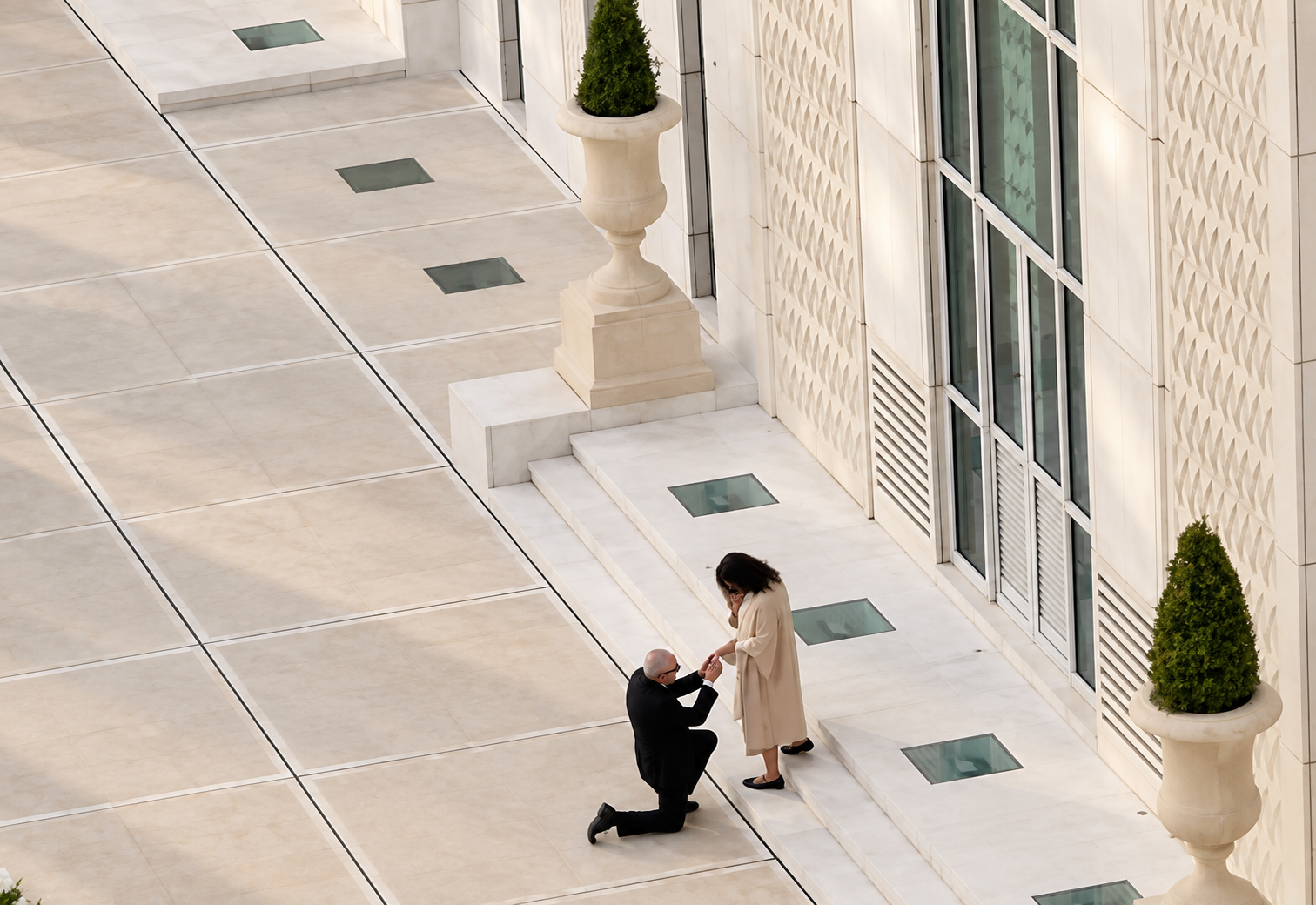 Una persona se arrodilla para proponerle matrimonio a otra en una luminosa terraza de piedra junto a un edificio.