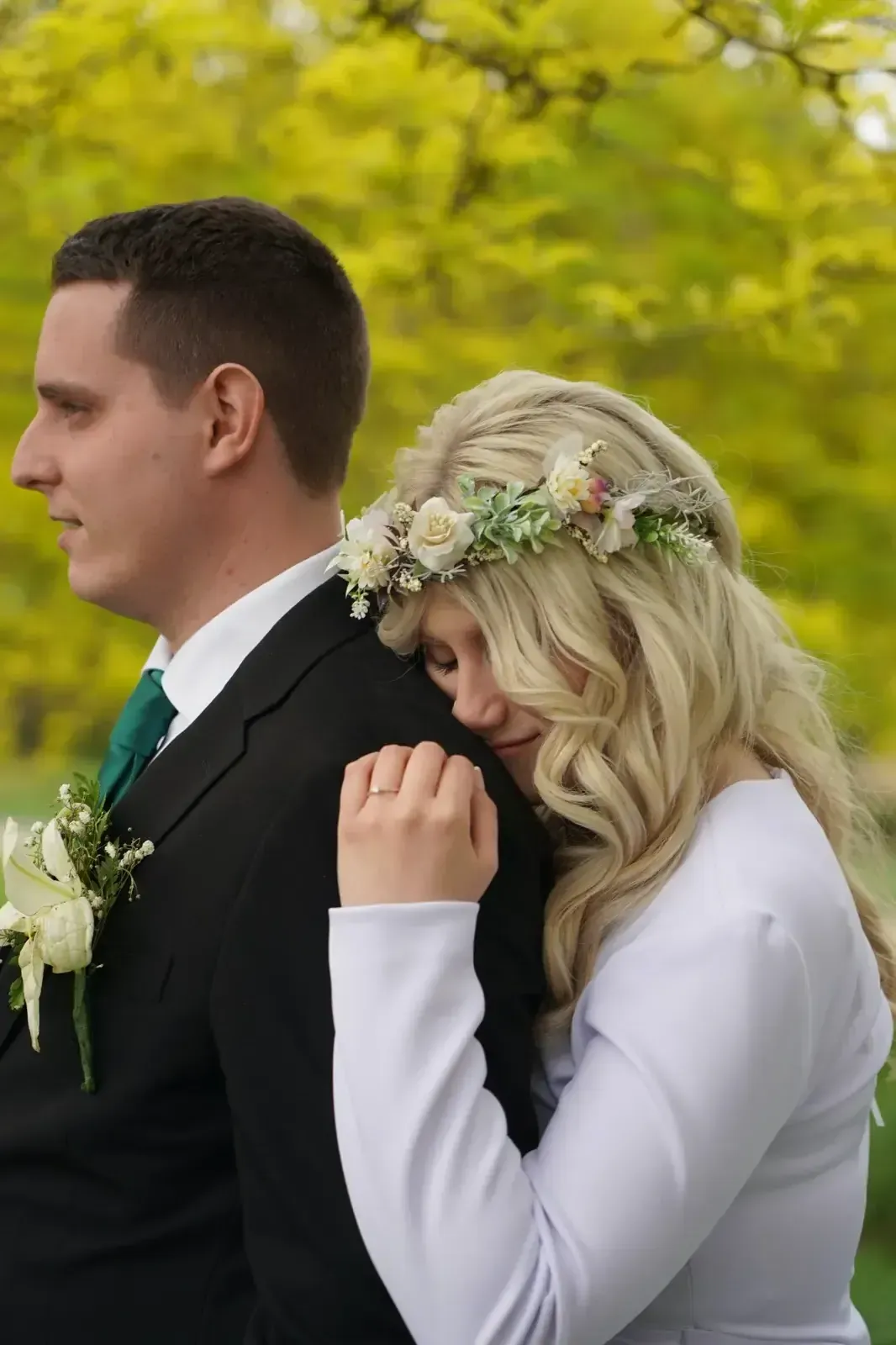La novia apoyada en el hombro del novio al aire libre, ambos con atuendo formal con detalles florales, sobre un fondo verde.
