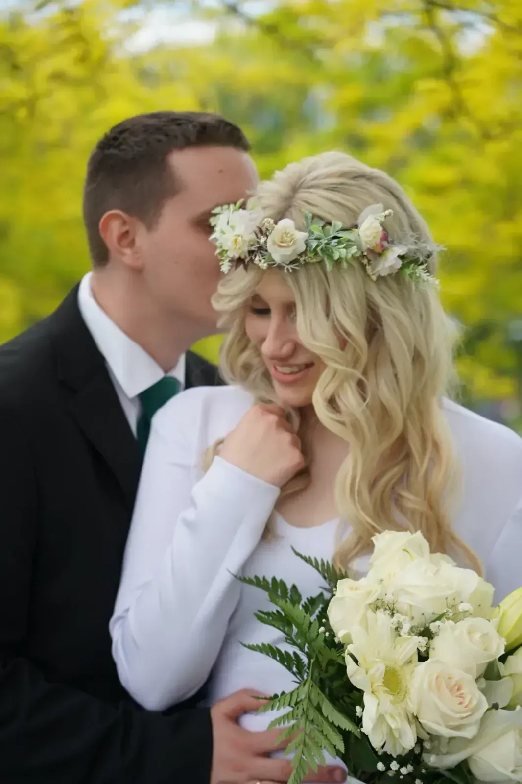 Pareja vestida de novios abrazándose al aire libre, con ramo blanco y corona de flores.