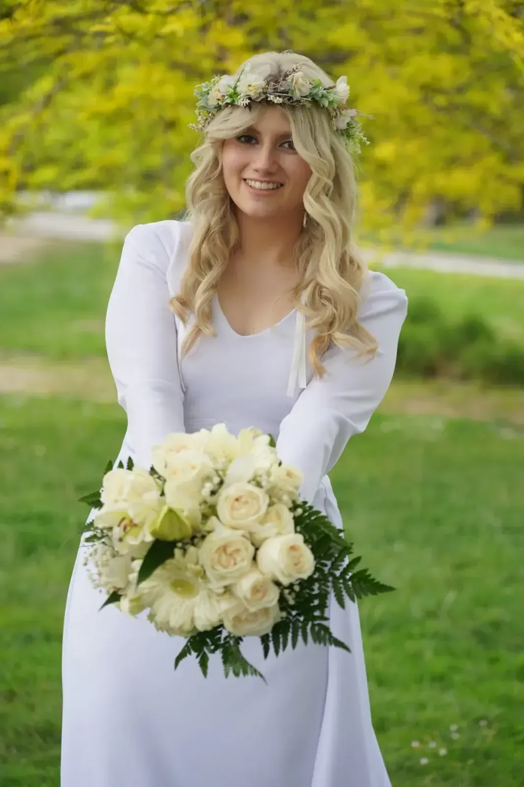 Una persona sonriente vestida de blanco sostiene un ramo de rosas color crema al aire libre con una corona de flores.
