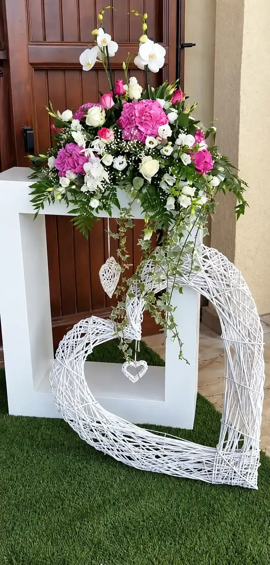Adorno de mimbre blanco en forma de corazón con flores rosas y blancas sobre un soporte sobre césped verde.