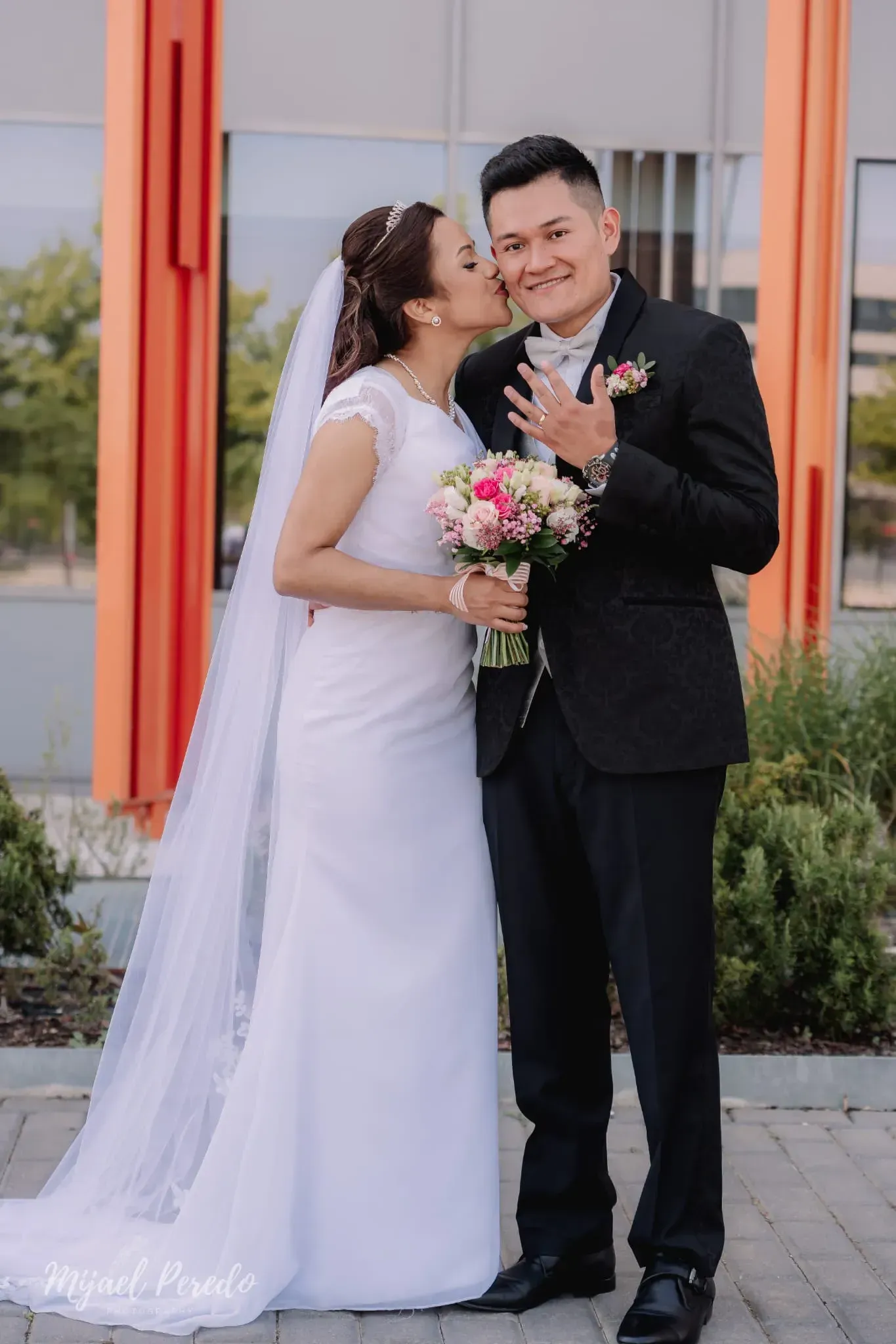 La novia besa al novio en la mejilla, ambos vestidos de traje de boda, de pie al aire libre junto a columnas naranjas.