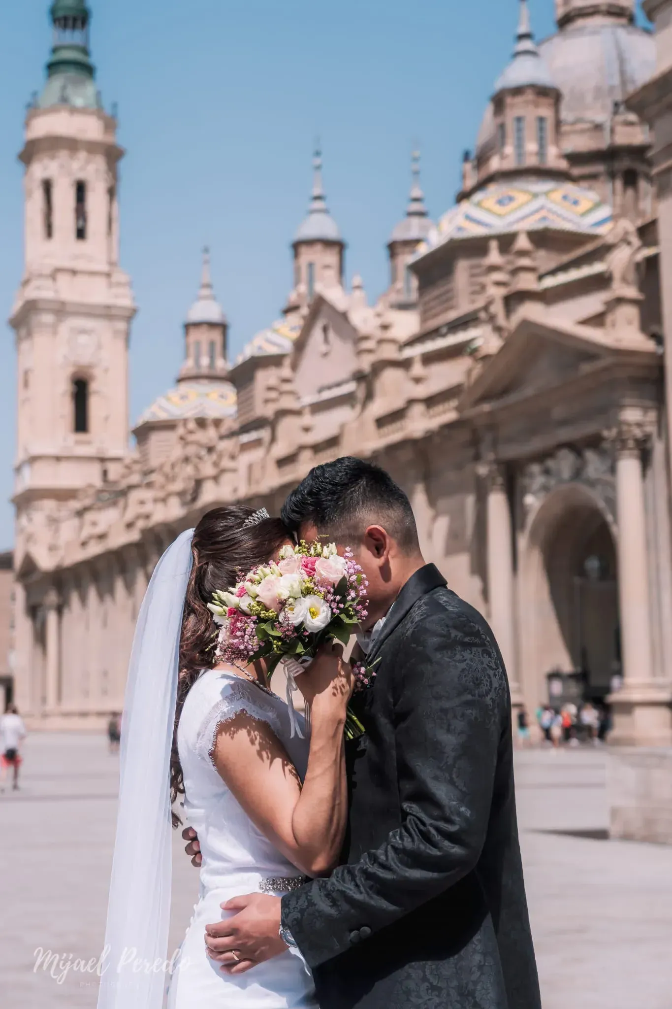 Pareja abrazándose con atuendo nupcial frente a una ornamentada fuente de la catedral.