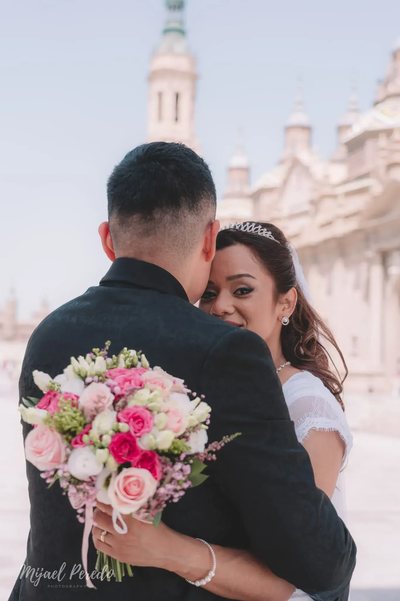 Los novios se abrazan, sosteniendo un ramo de flores rosas y blancas en el luminoso patio de una iglesia.