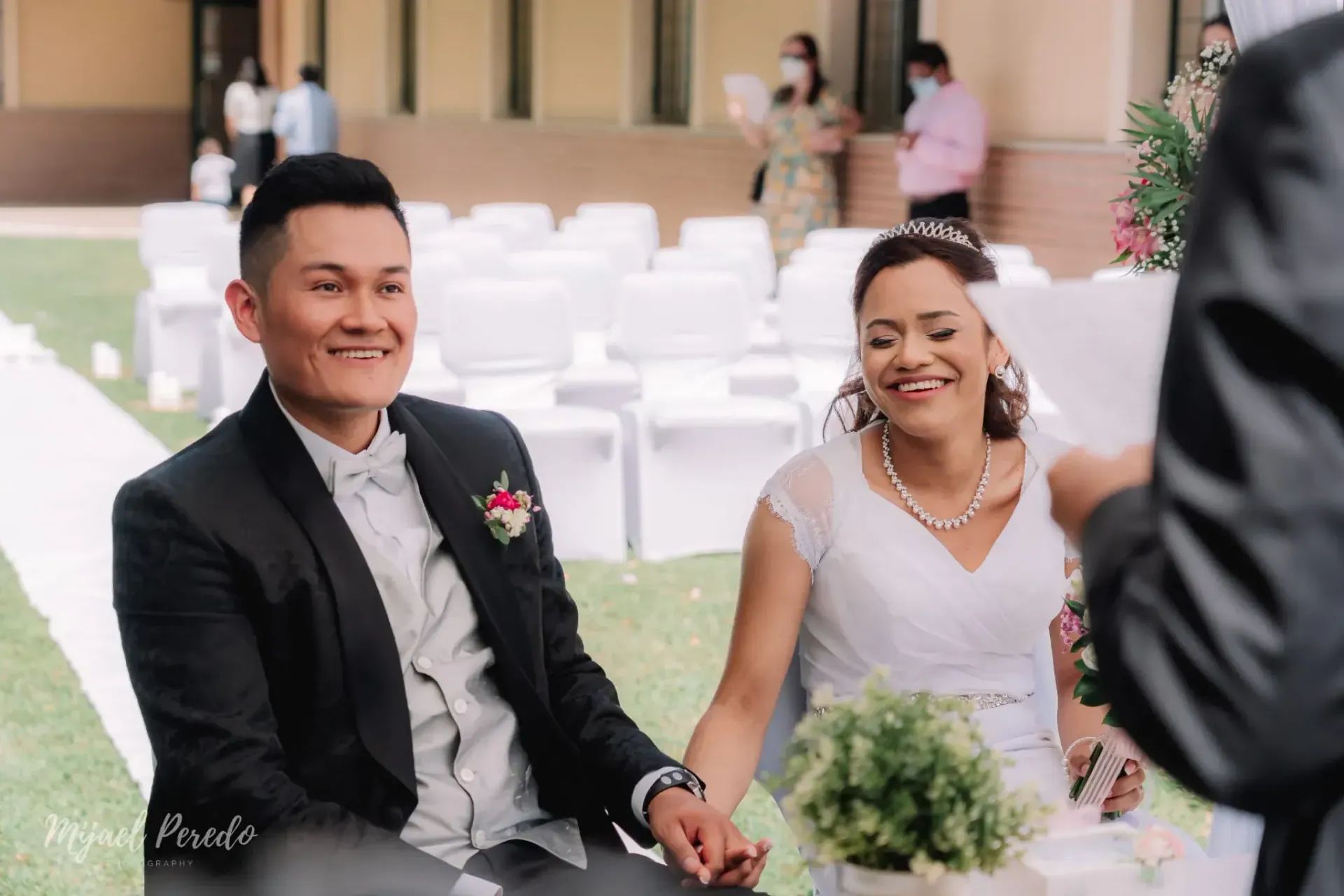 Los novios, sentados al aire libre, sonríen y se toman de la mano durante la ceremonia nupcial.