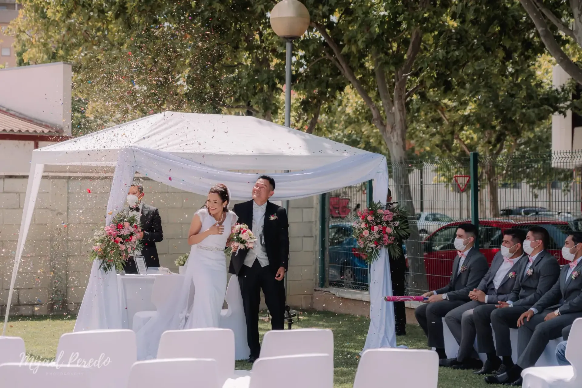 Ceremonia nupcial al aire libre bajo un dosel blanco con los novios de pie en el altar, los invitados sentados cerca.