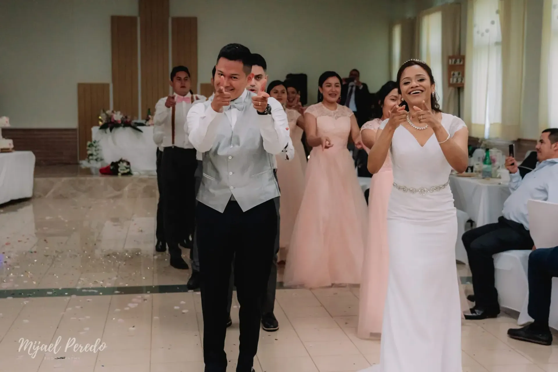 Los invitados a la boda caminan por el pasillo en un salón decorado, lanzando besos a la cámara.
