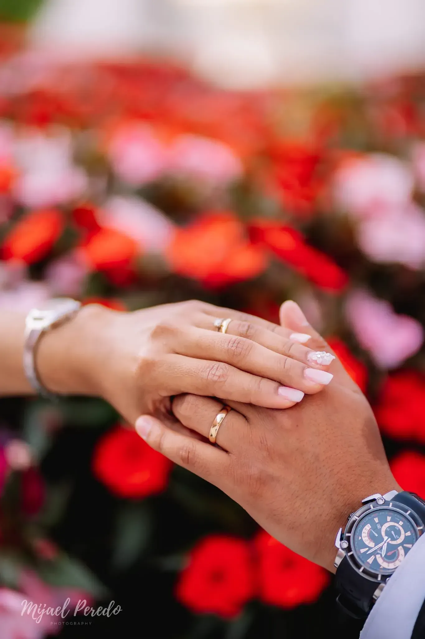Manos entrelazadas con un anillo y un reloj, sobre un fondo borroso de flores rojas.