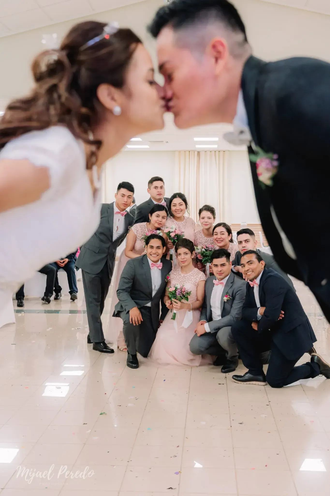 Grupo de novios posando en el pasillo, con la pareja besándose en primer plano y los invitados sonriendo detrás.