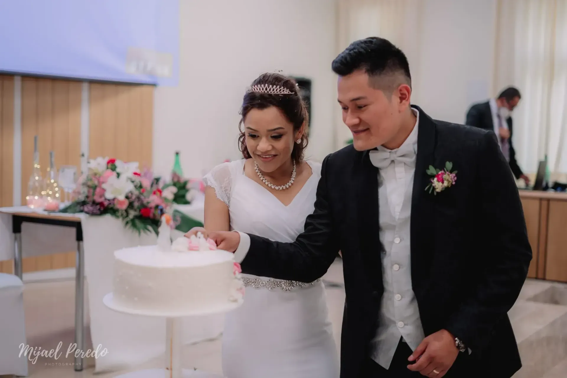 Los novios cortan un pastel de bodas blanco y sonríen en la mesa de la recepción.