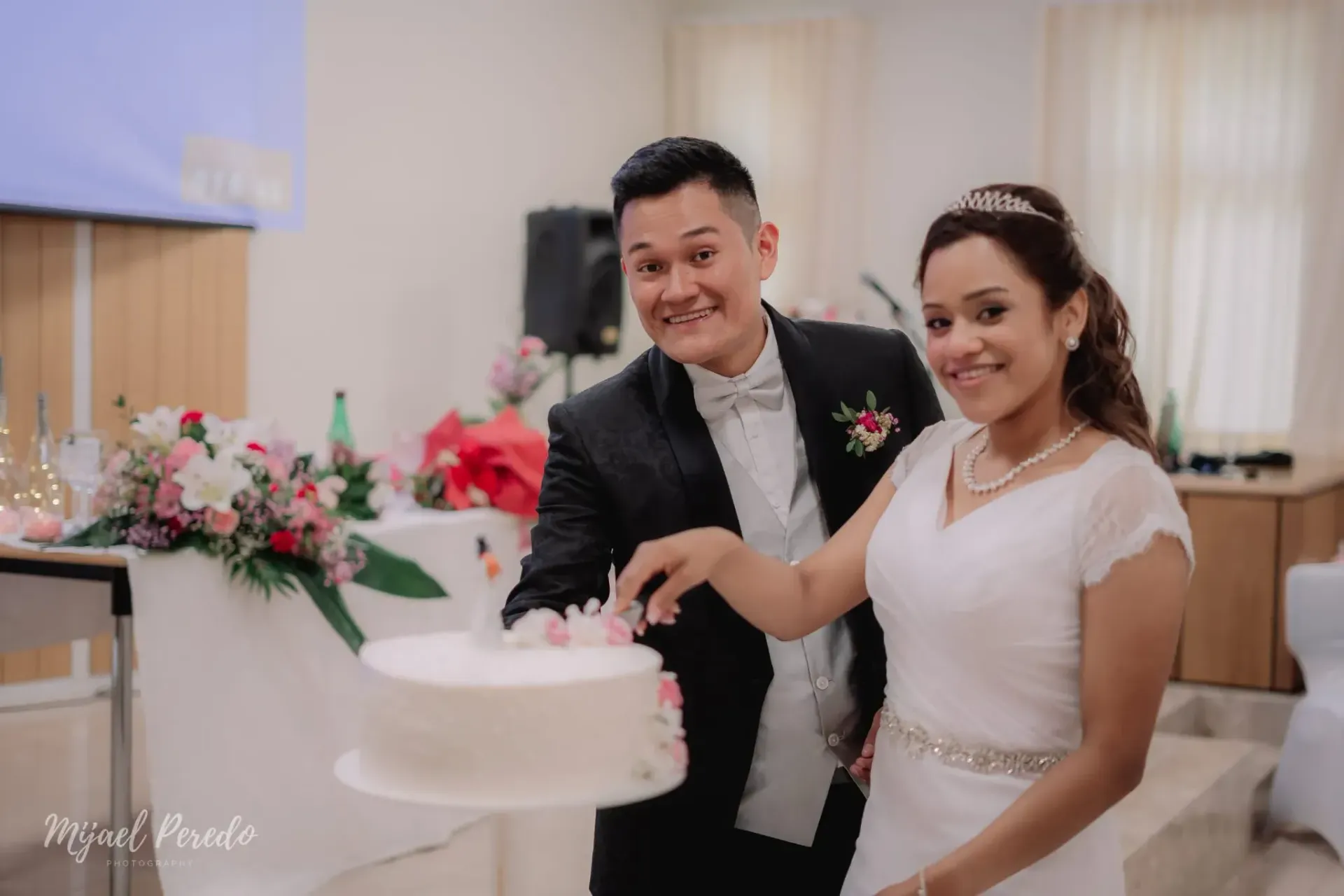 Los novios sonríen junto a un pastel de bodas blanco en su recepción.