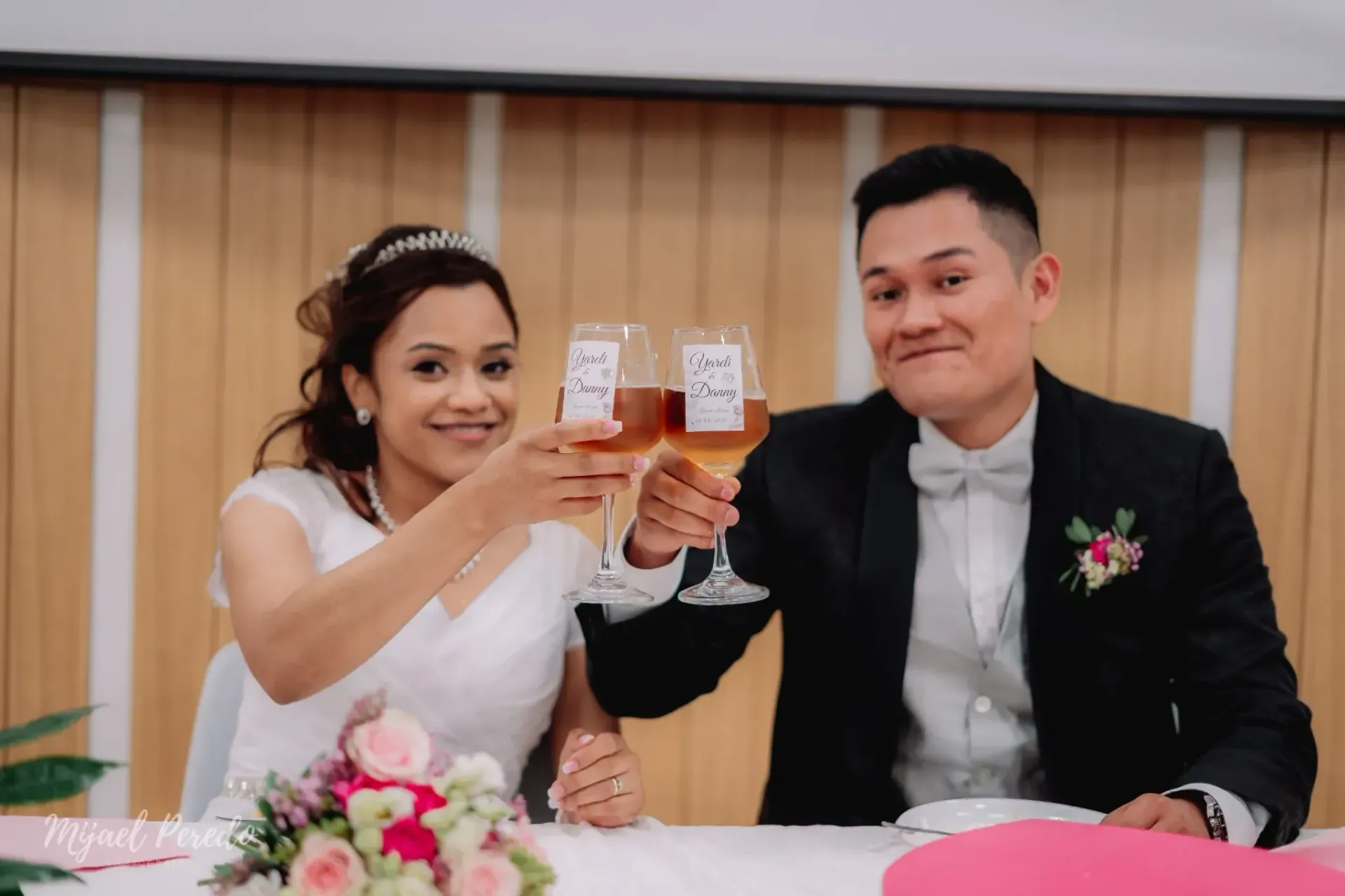 Una pareja vestida de novios brinda con bebidas en una mesa decorada, sonriendo frente a un panel de madera.