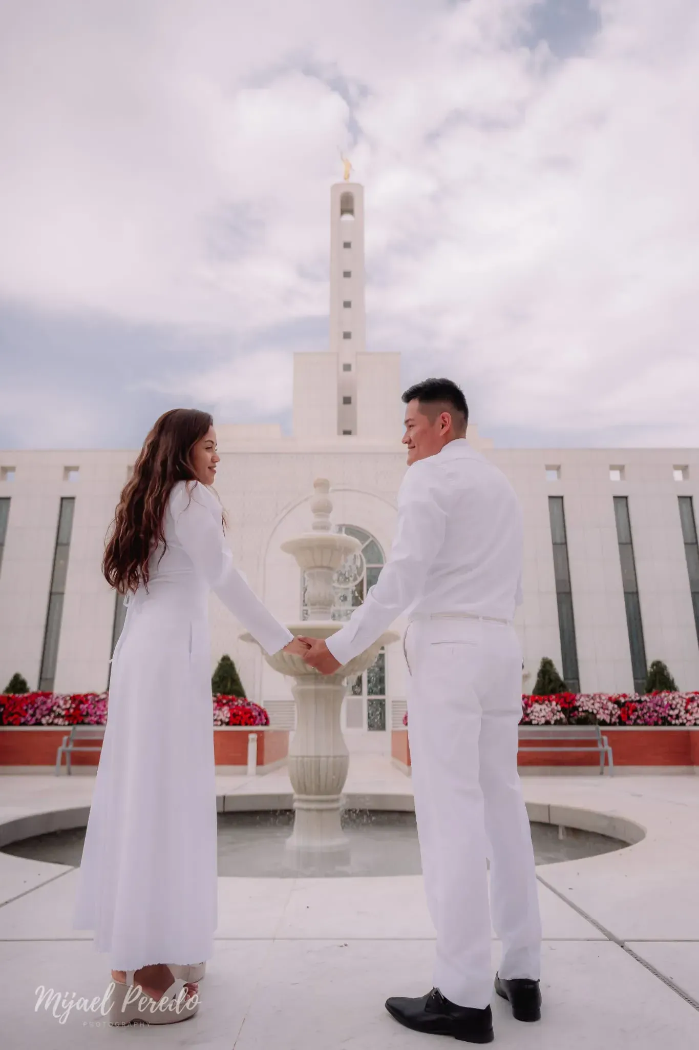 Una pareja vestida de blanco, tomada de la mano, se encuentra frente a un alto monumento blanco en un patio luminoso.