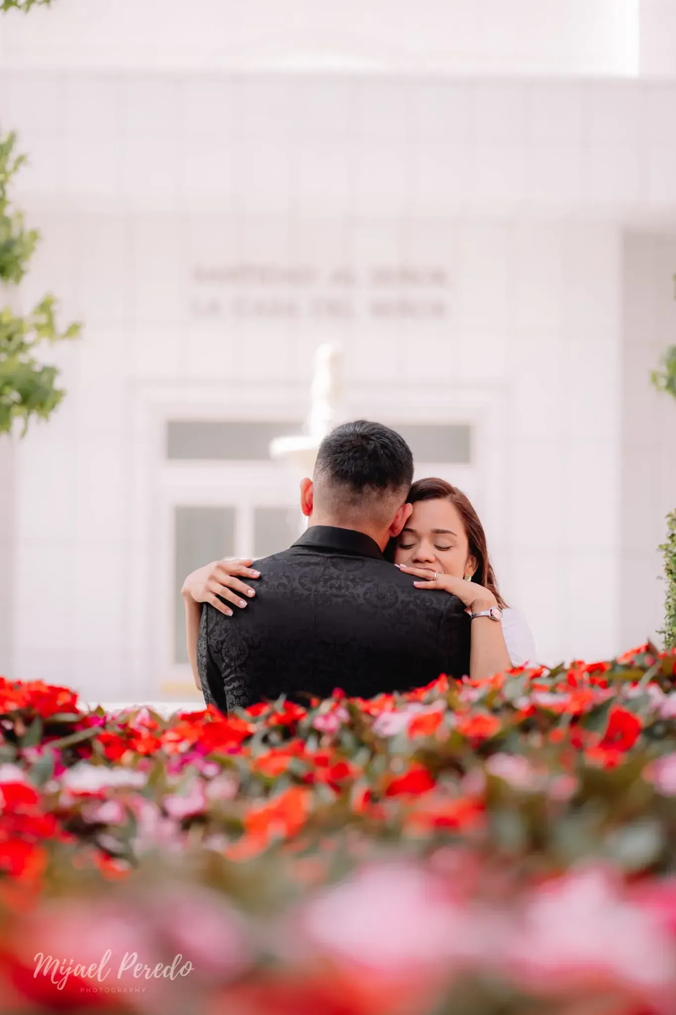 Una pareja se abraza en un jardín blanco, con flores rojas en primer plano.