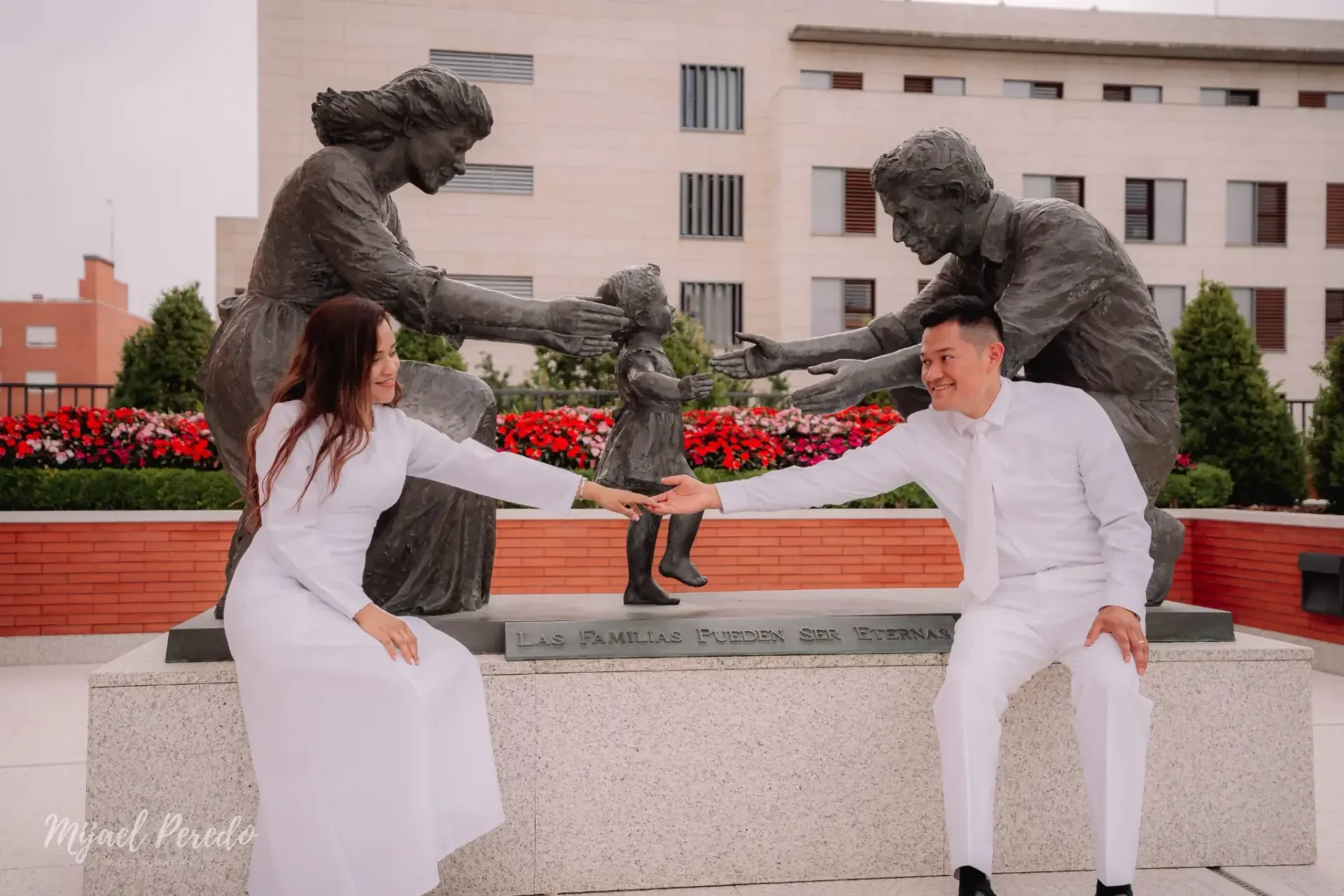 Dos personas vestidas de blanco posan con un perro pequeño en un banco frente a una estatua de bronce al aire libre.