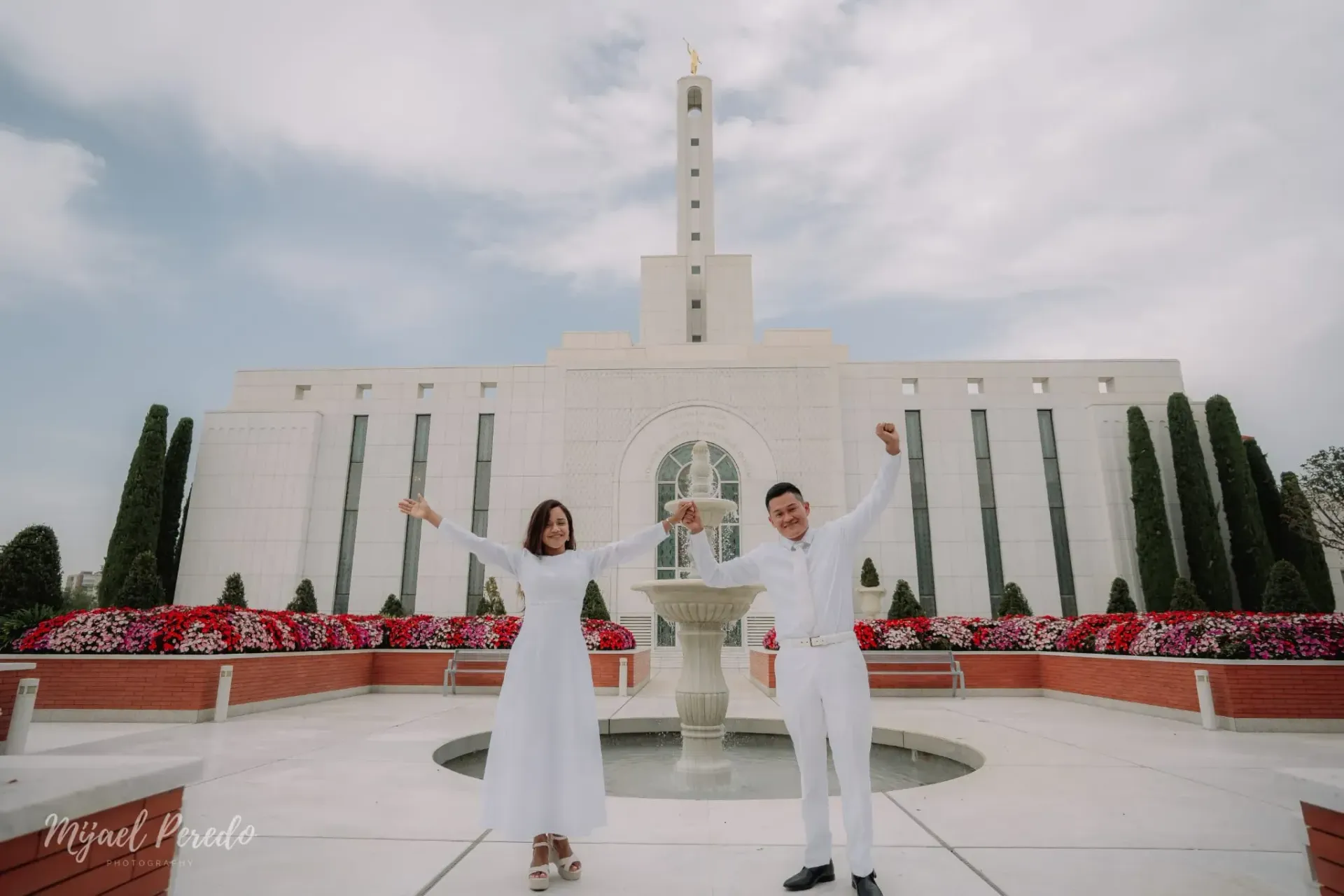 Dos personas vestidas de blanco se encuentran de pie frente a un gran templo blanco con una fuente y flores rojas.