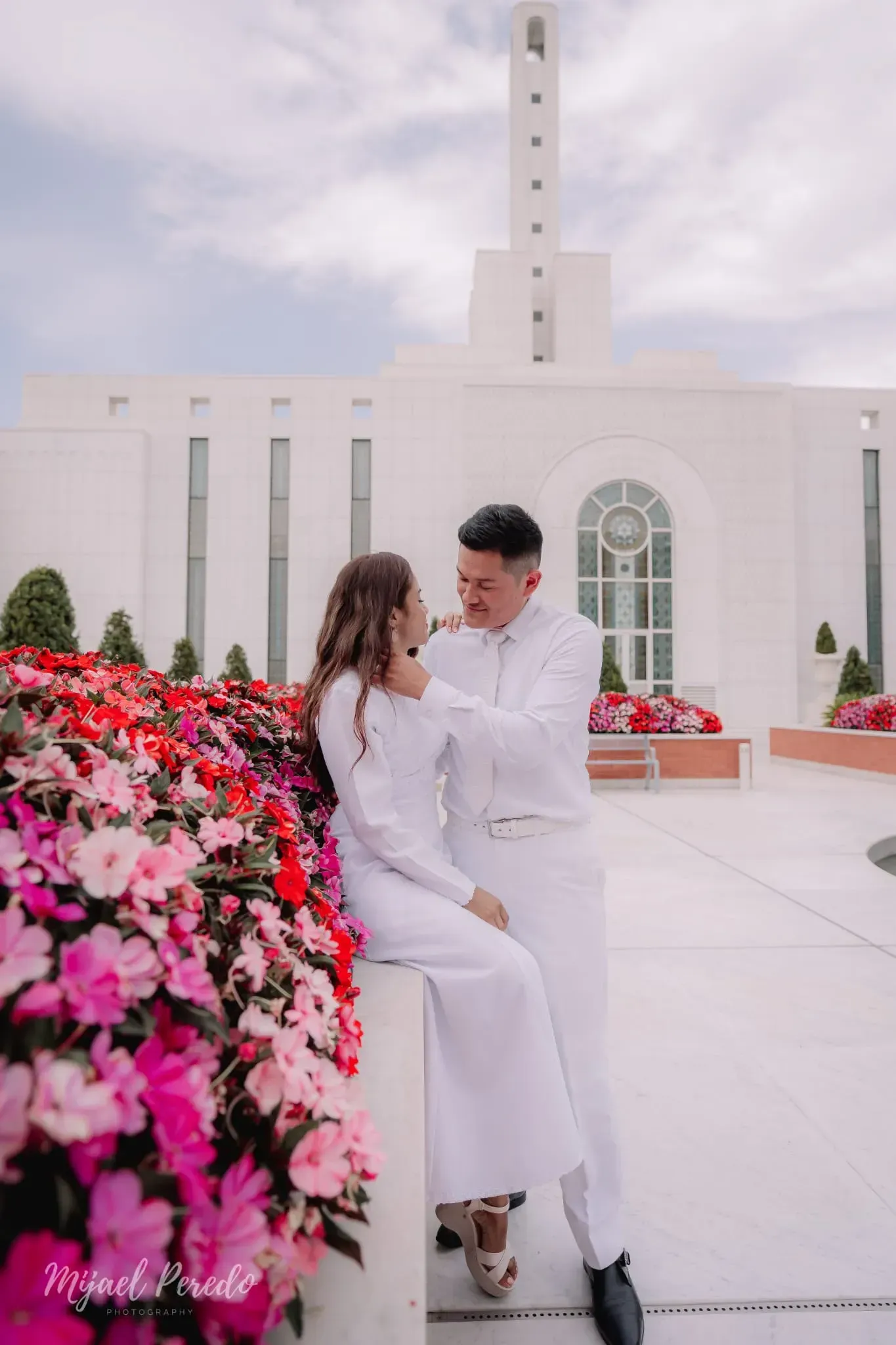 Una pareja vestida de blanco sentada junto a flores de color rosa brillante