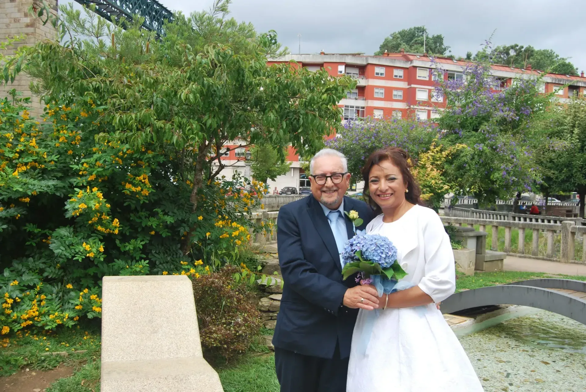 Pareja con atuendo formal sosteniendo flores junto a un puente de jardín con edificios al fondo.