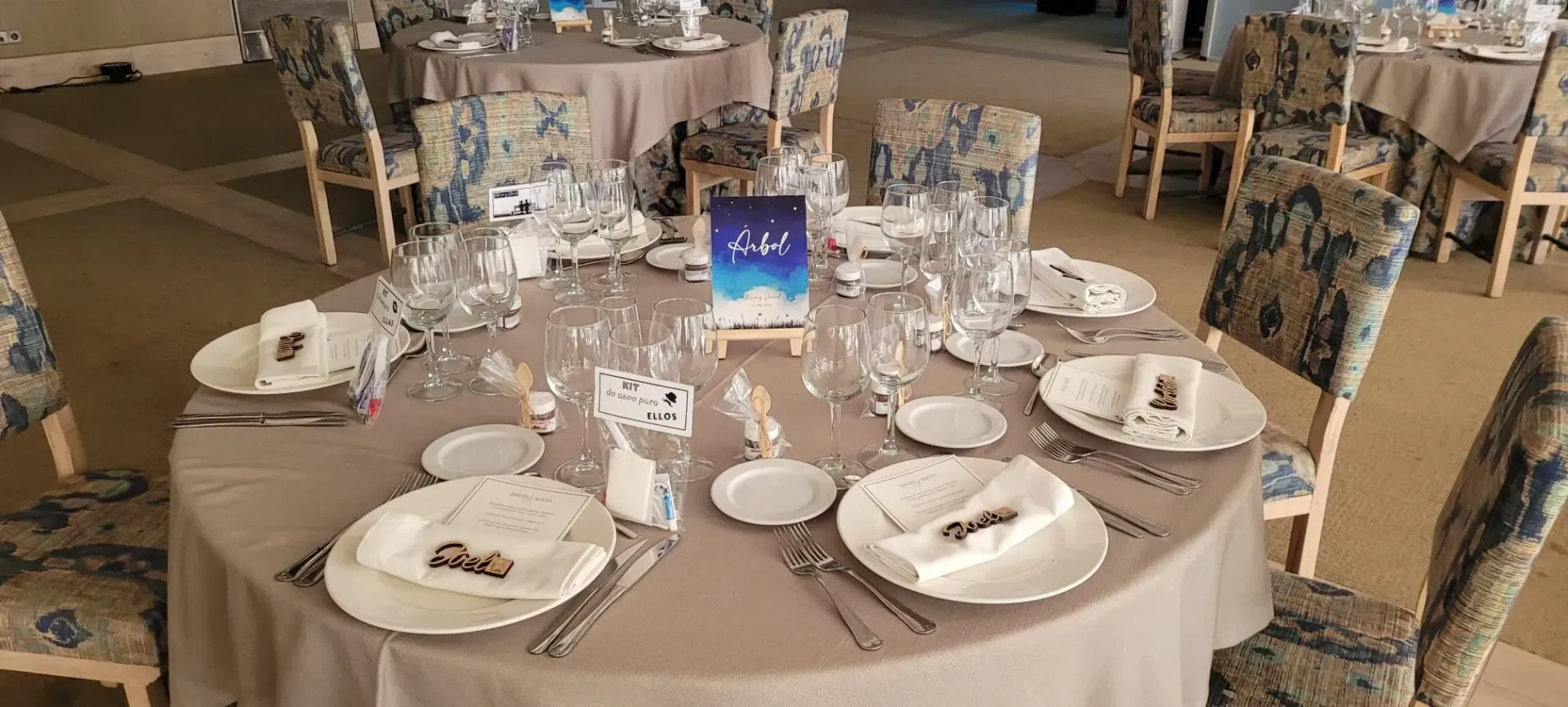 Mesa redonda de banquete puesta con platos blancos, vasos y un centro de mesa azul-púrpura en un lugar para eventos.