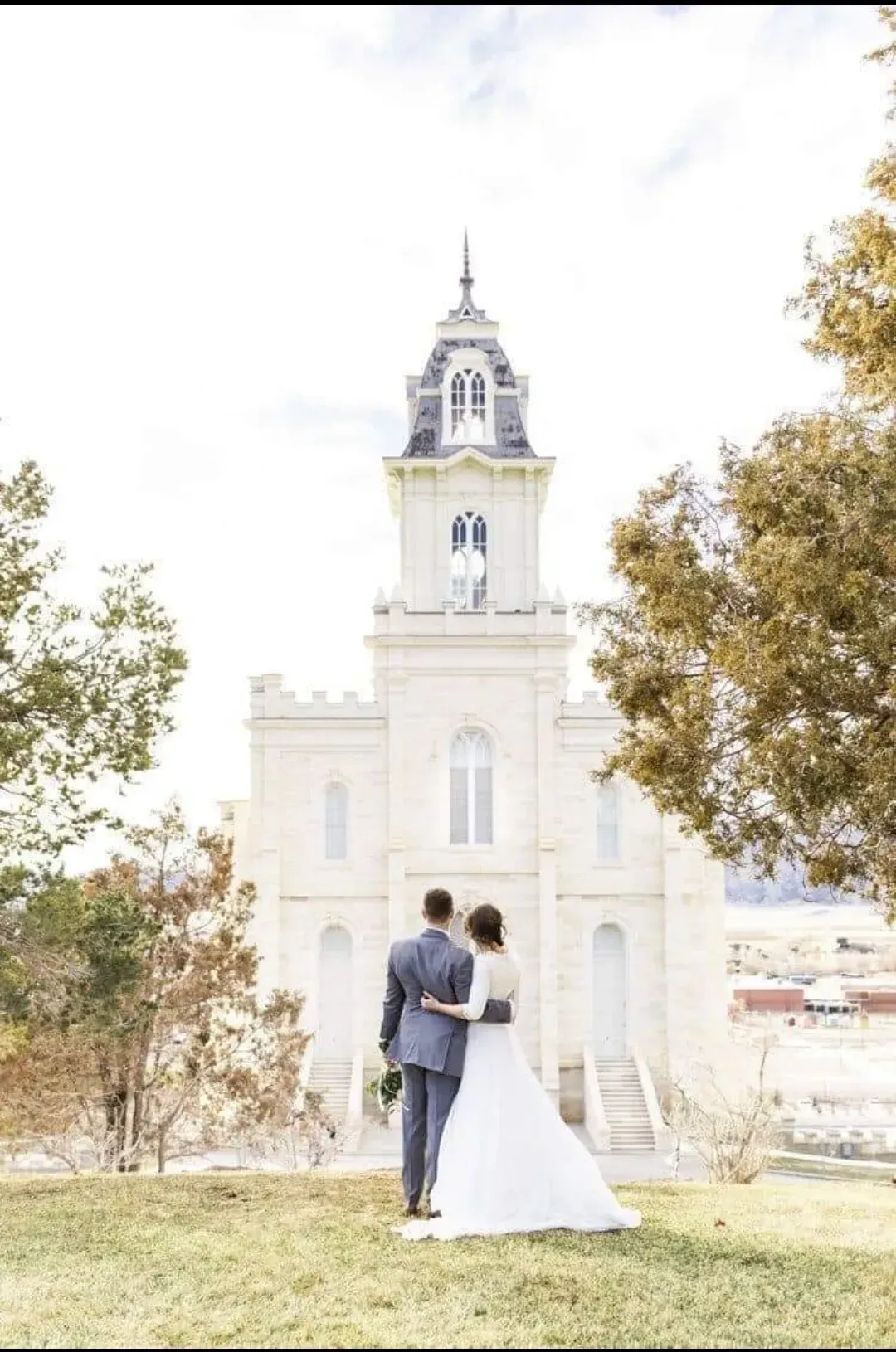 Una pareja vestida de novios posa frente a la torre blanca de una iglesia histórica al aire libre.