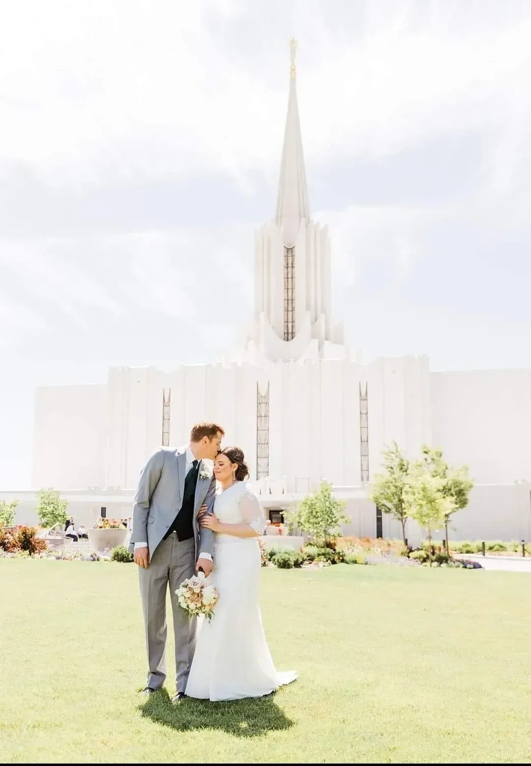 Una pareja vestida de boda se abraza sobre un césped frente a un edificio blanco con aspecto de templo.