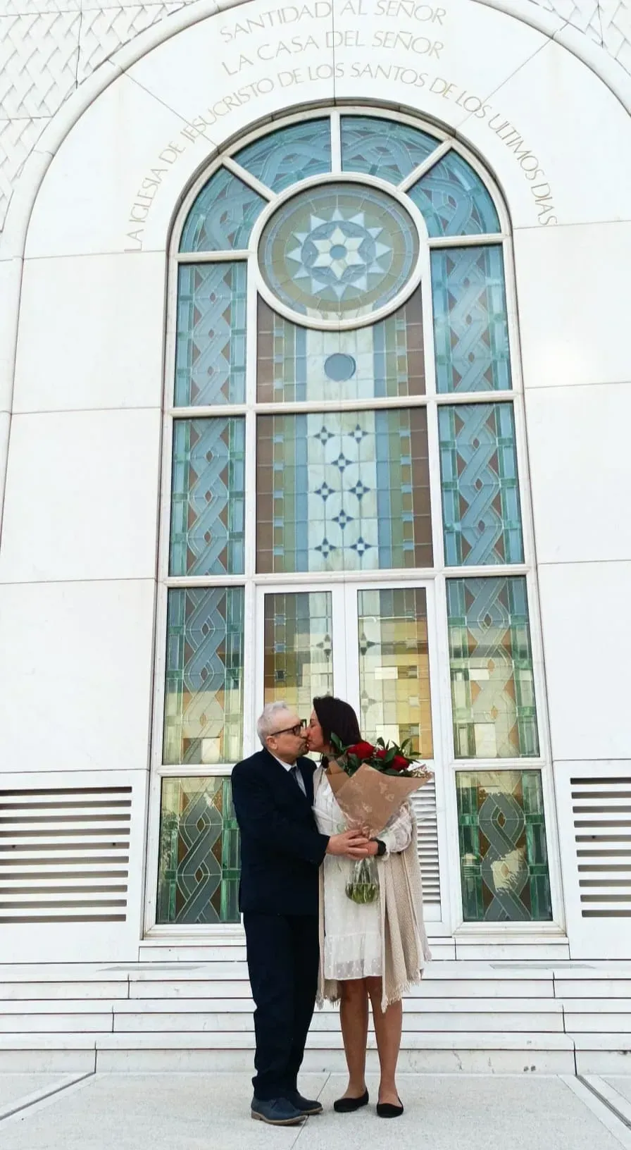 Una pareja se abraza vestida de boda frente a una alta ventana arqueada en un edificio blanco.