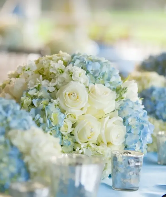 Ramos de rosas blancas y azul pálido en jarrones de plata sobre una mesa decorada.