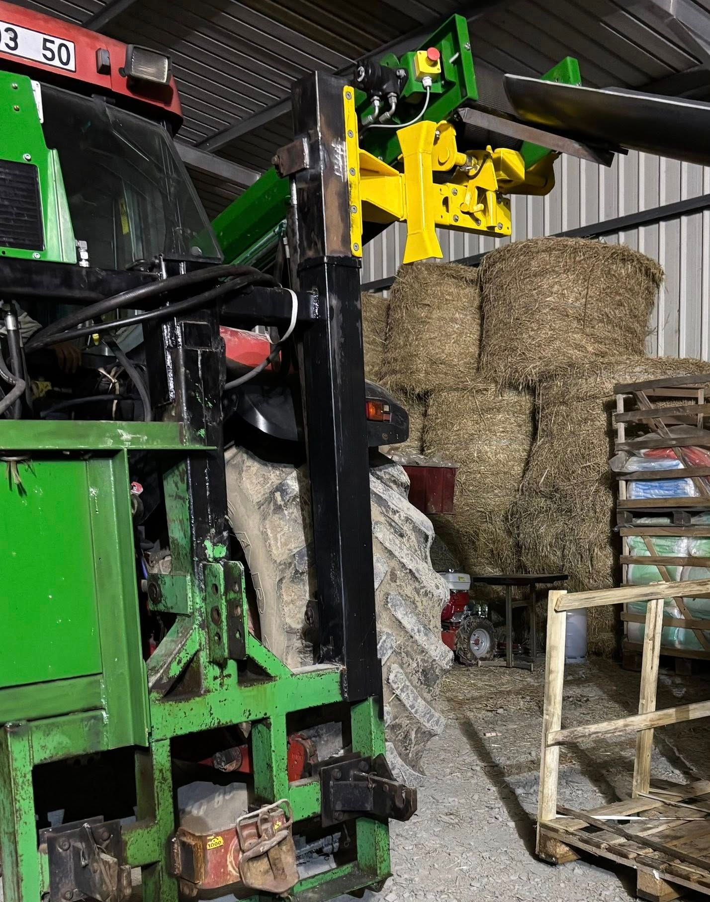 Une machine agricole avec un bras en métal, des bottes de foin sont empilées en arrière-plan.