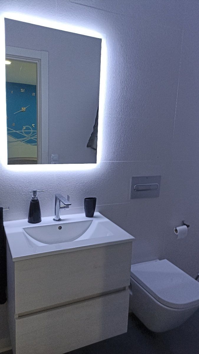 Baño con paredes blancas, espejo iluminado, lavabo blanco, inodoro y dispensador de jabón.