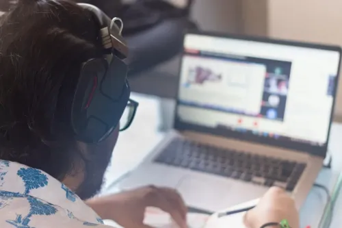 Persona con auriculares y gafas, participando en una reunión virtual desde un ordenador portátil; tomando notas.