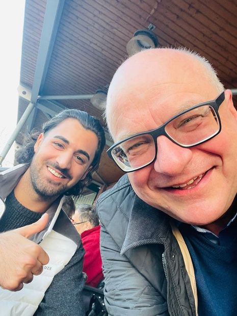 Zwei Männer lächeln, einer zeigt den Daumen nach oben. Szene im Freien, ein rotes Hemd ist teilweise sichtbar.