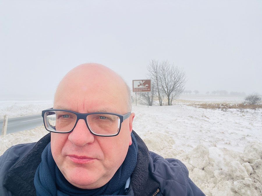 Glatzköpfiger Mann mit Brille in verschneiter Landschaft, im Hintergrund eine Straße und ein Schild.