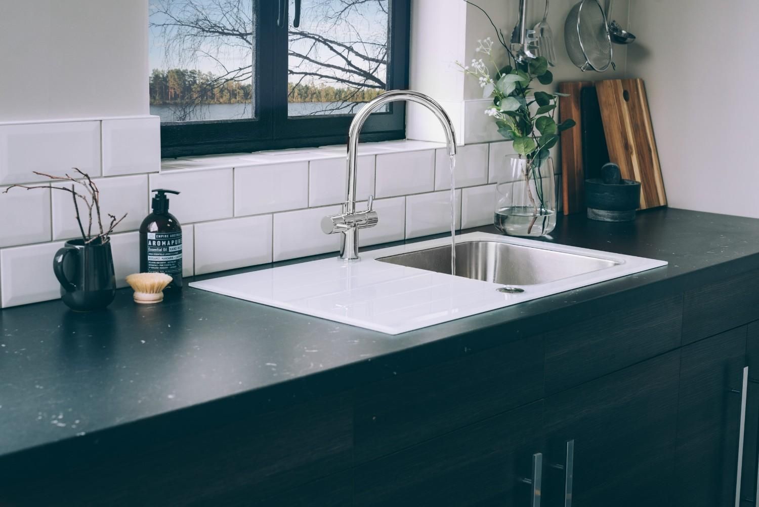 Fregadero de cocina moderno con encimera negra, salpicadero de azulejos blancos, ventana y pequeños elementos decorativos.