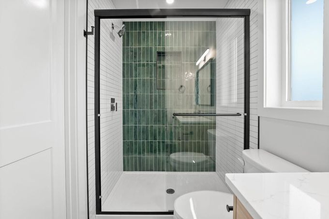 Baño moderno con ducha de cristal, puerta con marco negro, lavabo blanco y paredes de azulejos verdes.