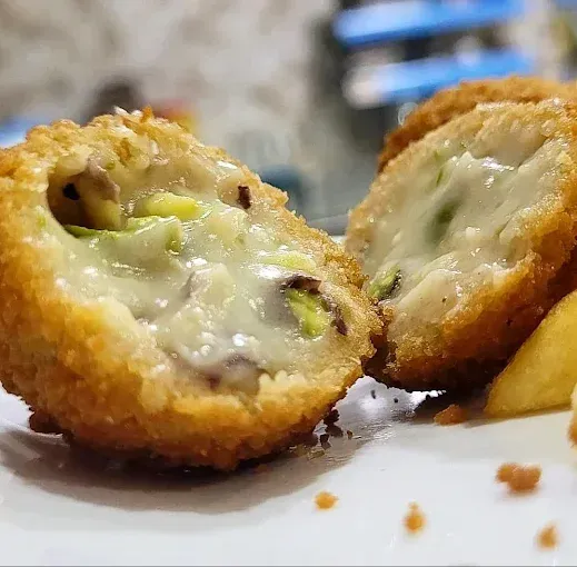 Primer plano de un crujiente aperitivo frito cortado por la mitad, que deja ver el relleno cremoso con trocitos de pistacho.