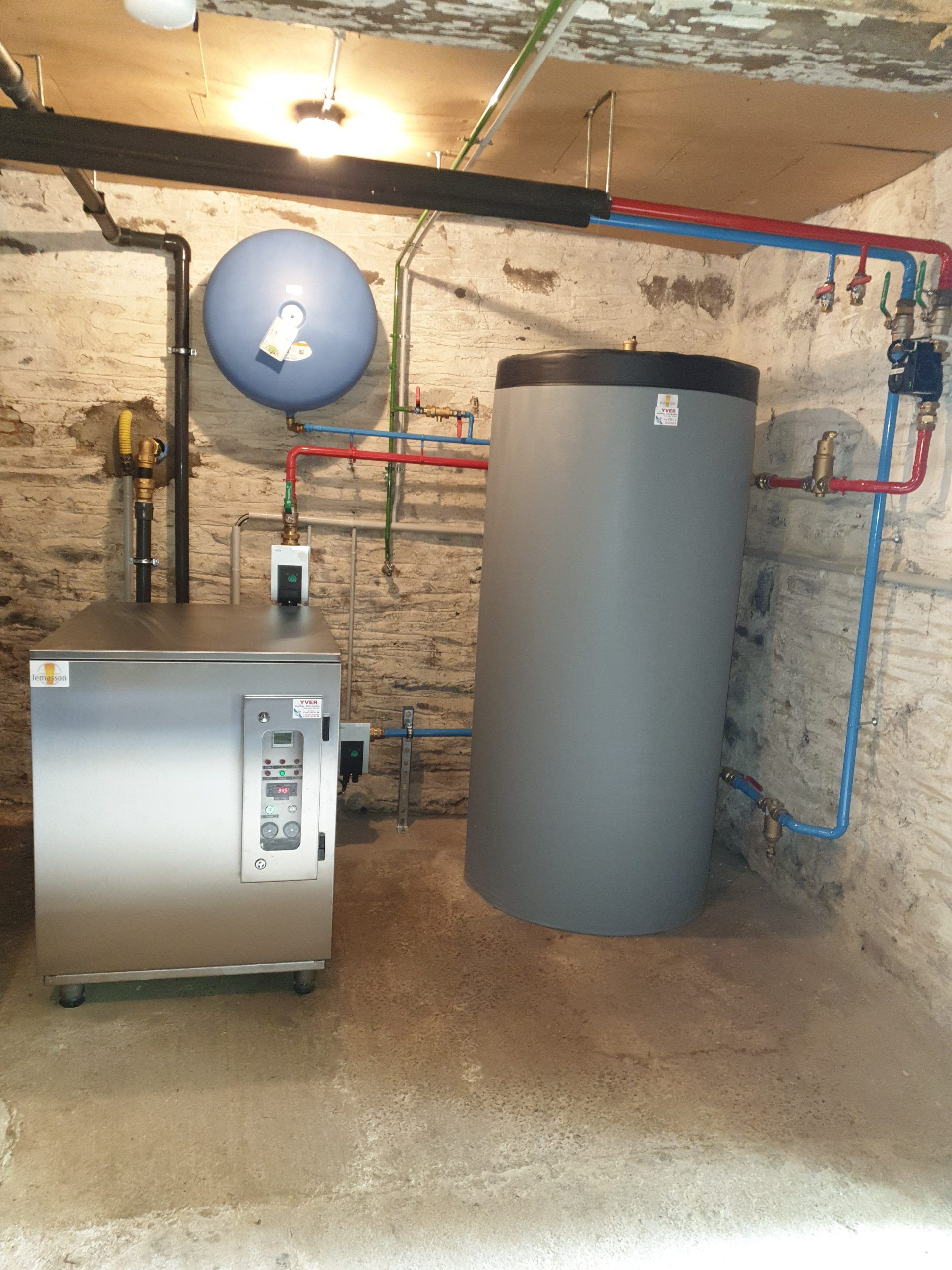 Local technique au sous-sol avec chaudière, réservoir d'eau, tuyauterie et vase d'expansion bleu fixé au mur.