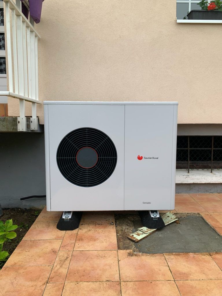 Unité de pompe à chaleur extérieure installée à côté d'une maison, avec un grand ventilateur noir et un boîtier blanc sur une terrasse.