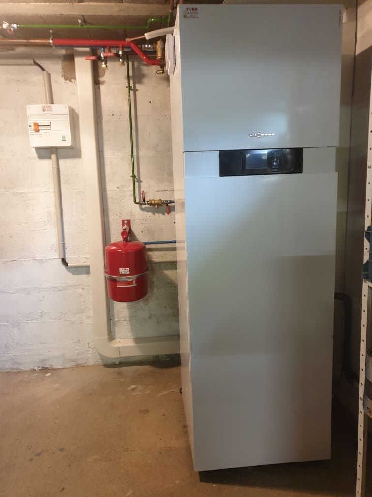 Local technique au sous-sol avec une grande unité de climatisation grise, un extincteur et des tuyaux apparents sur un mur en béton.